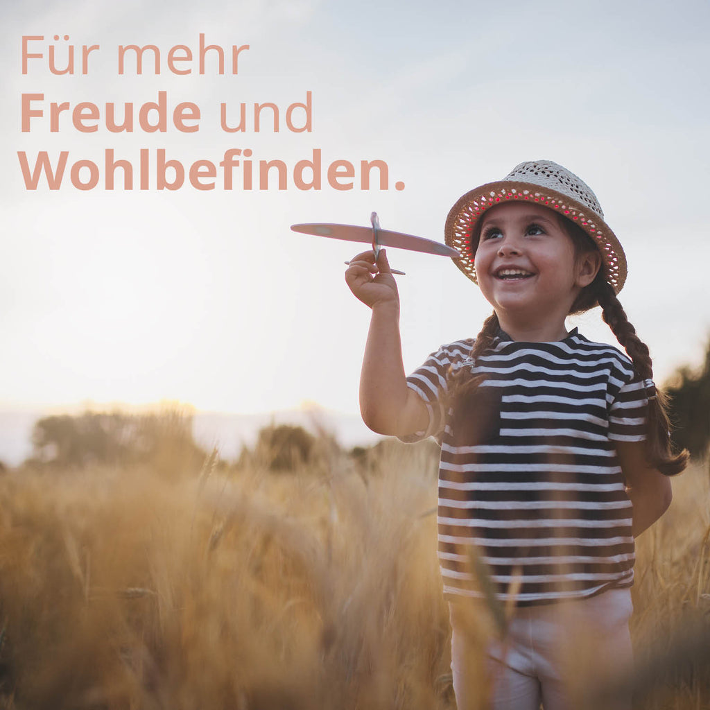 Ein kleines Mädchen mit geflochtenen Zöpfen, Hut und gestreiftem Oberteil hält ein Modellflugzeug in der rechten Hand und lächelt. Text: Für mehr Freude und Wohlbefinden. 