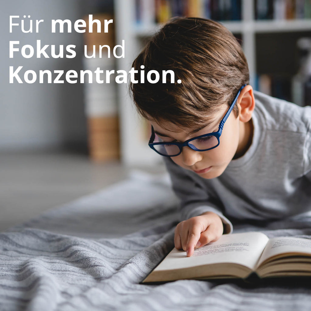 Kleiner Junge mit blauer Brille liegt auf dem Bett und beugt sich zu einem Buch vor - er wirkt konzentriert. Text: Für mehr Fokus und Konzentration