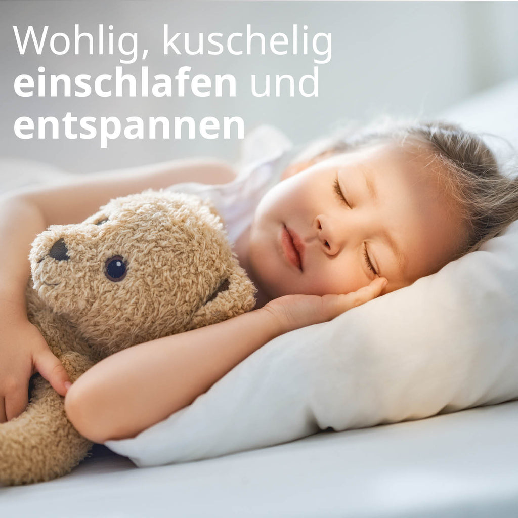 Ein kleines Mädchen liegt seitlich im Bett, hält einen Teddybären im Arm und schläft. Text: Wohlig, kuschelig einschlafen und entspannen